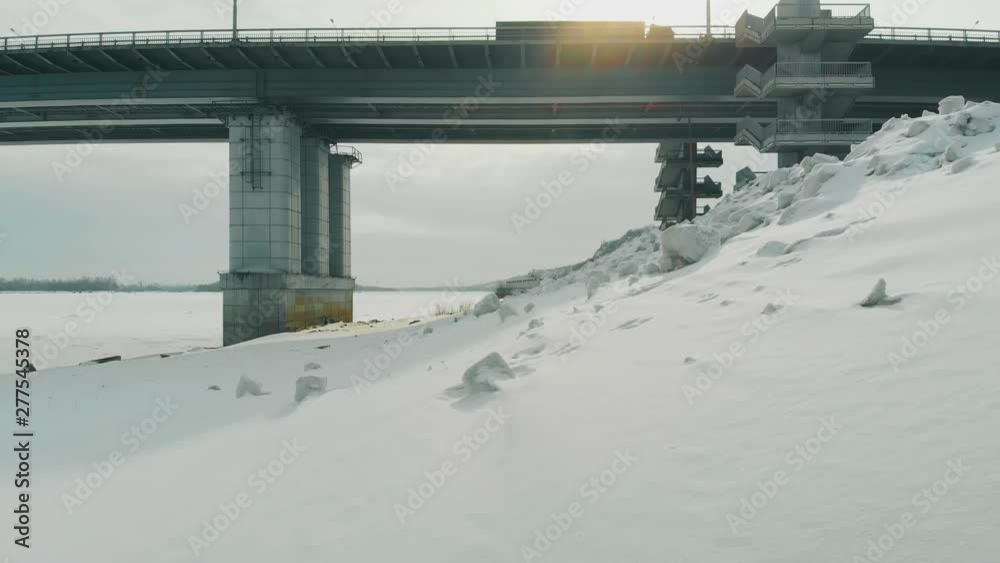 trucks drive along huge bridge near old rusty pier on wide frozen river bank covered with snow upper view trucks drive along huge bridge near old rusty pier on wide frozen river bank covered with snow upper view