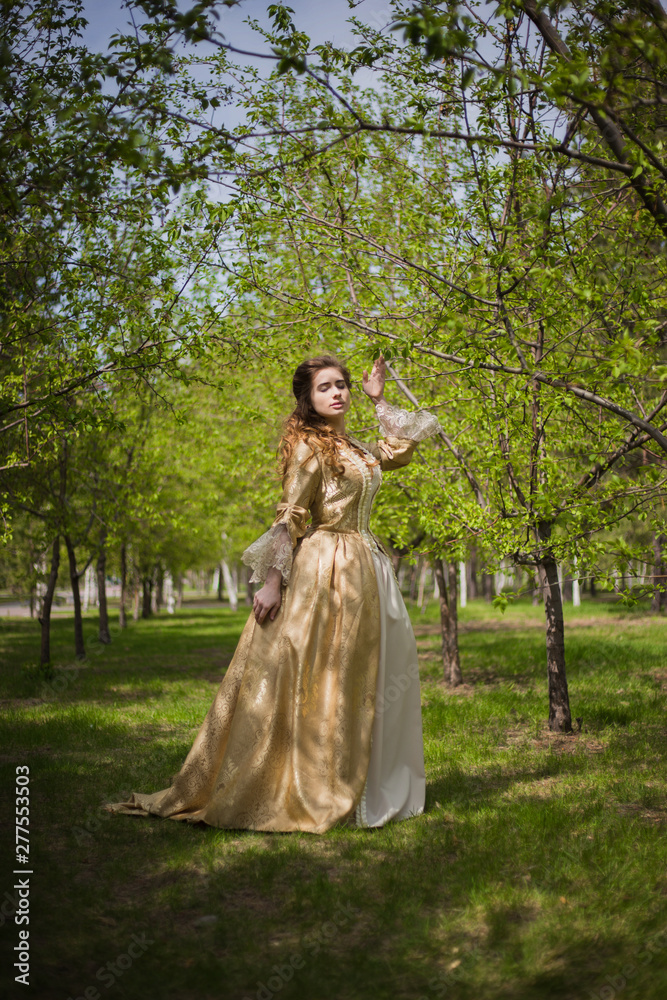 Beautiful girl in a gold dress against a background of green trees. A woman in a ball gown of the Rococo era.