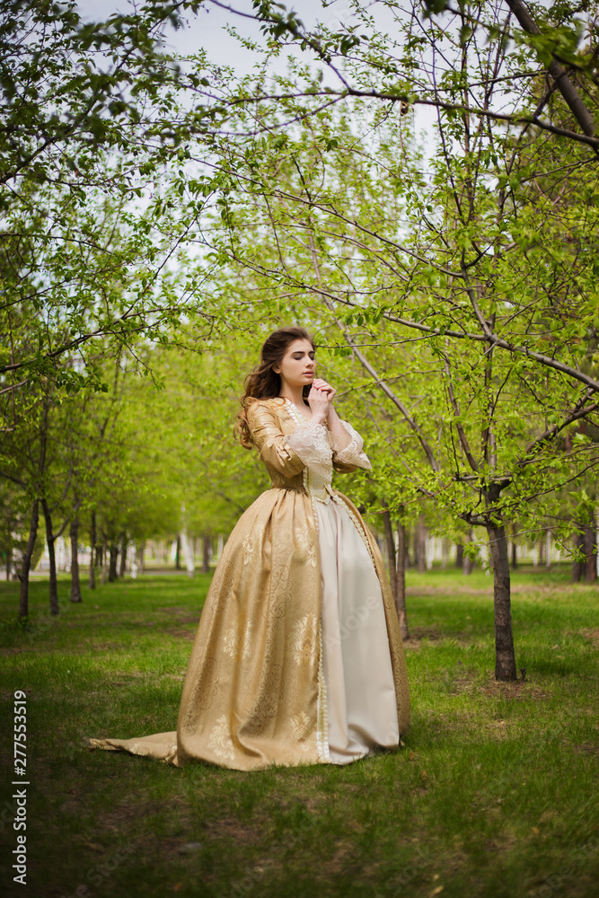 Beautiful girl in a gold dress against a background of green trees. A woman in a ball gown of the Rococo era. Beautiful girl in a gold dress against a background of green trees. A woman in a ball gown of the Rococo era.