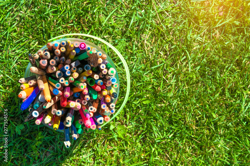 a bunch of colored pencils in a plastic bucket on the green grass sunbeam in the upper right corner top view