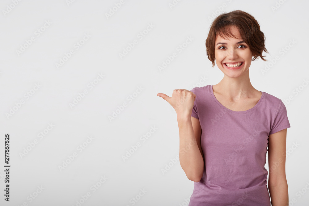 Cute cheerful short haired young woman in blank t-shirt, lloks happy and broadly smiles, wonts to draw you attention and points fingers to the copy space on the left side over white wall. Cute cheerful short haired young woman in blank t-shirt, lloks happy and broadly smiles, wonts to draw you attention and points fingers to the copy space on the left side over white wall.