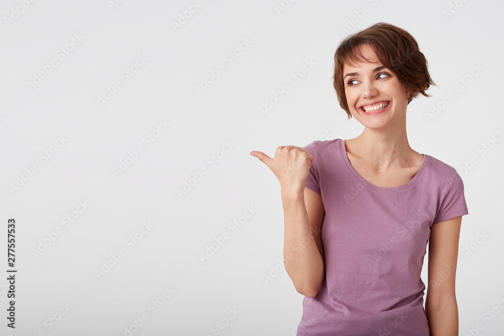 Photo of cute cheerful short haired young woman in blank t-shirt, lloks happy and broadly smiles, looks and points fingers to the copy space on the left side over white wall. Photo of cute cheerful short haired young woman in blank t-shirt, lloks happy and broadly smiles, looks and points fingers to the copy space on the left side over white wall.