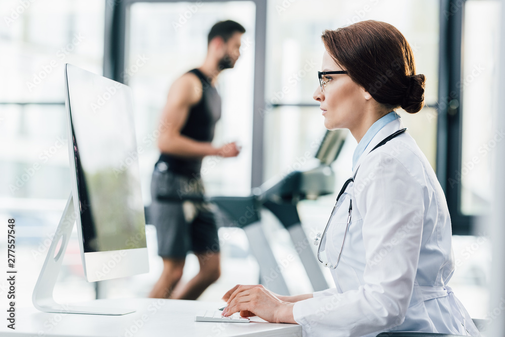 doctor in glasses using computer while sportsman running on treadmill during endurance test in gym
