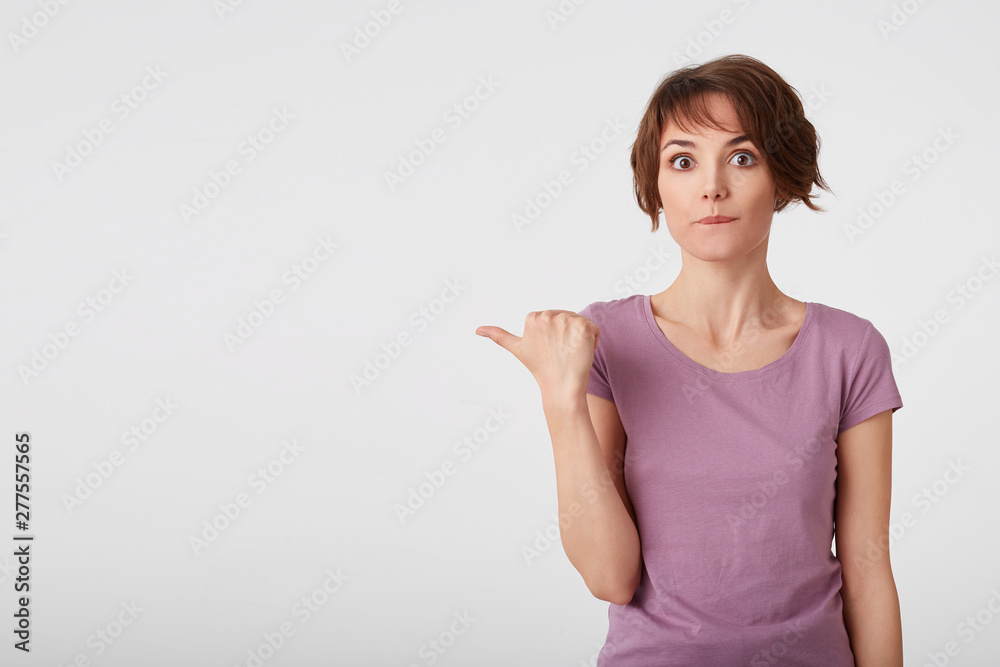 Portrait of confused nice short haired young woman in blank t-shirt, wonts to draw you attention and points fingers to the copy space on the left side over white wall. Portrait of confused nice short haired young woman in blank t-shirt, wonts to draw you attention and points fingers to the copy space on the left side over white wall.
