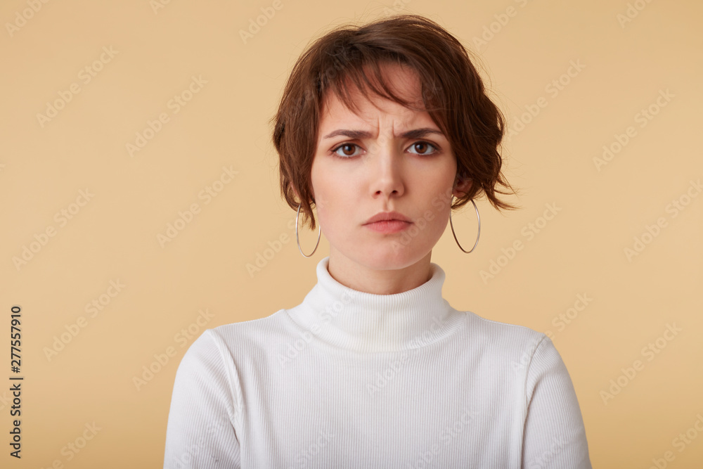 Close up of unhappy frowning short haired young lady in white golf, looks at the camera with sadly facial expression, stands over beige background. People and emotion concept. Close up of unhappy frowning short haired young lady in white golf, looks at the camera with sadly facial expression, stands over beige background. People and emotion concept.