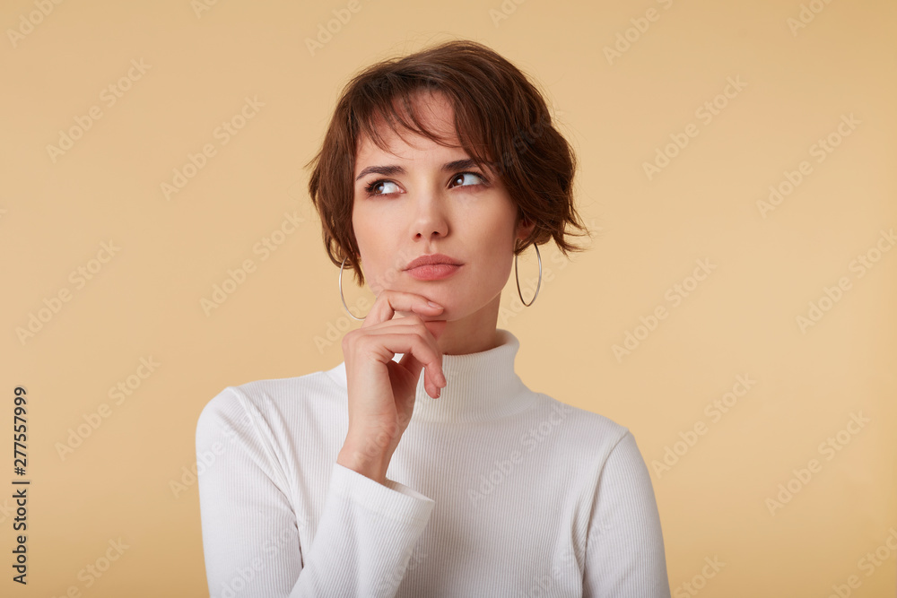 Close up of doubtful short haired young woman wears in white golf, stands over beige background, pensive looks away and touches chin.