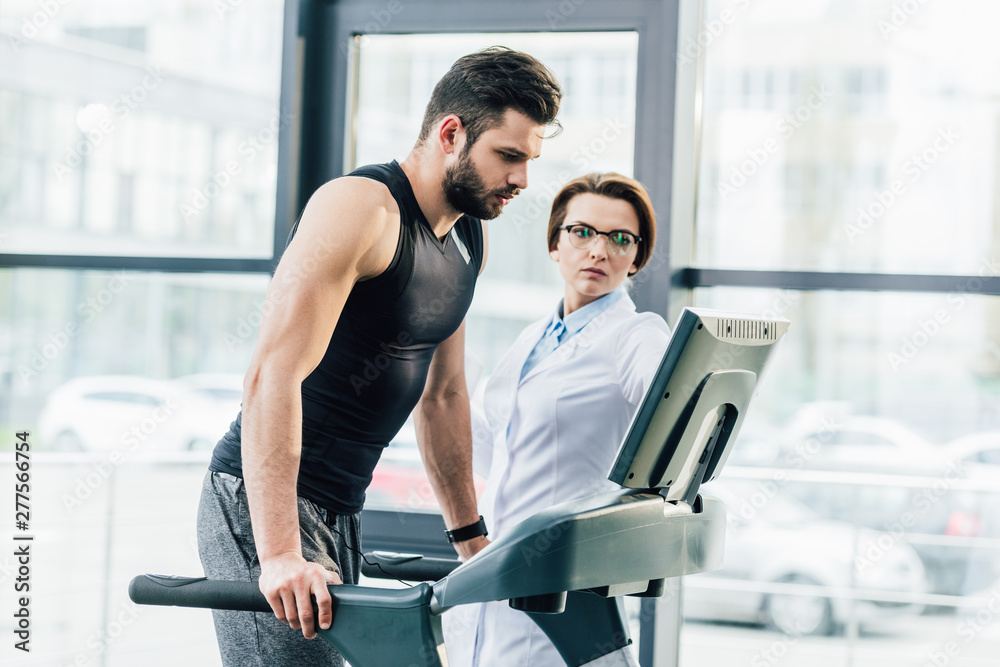 sportsman training on treadmill near doctor during endurance test in gym sportsman training on treadmill near doctor during endurance test in gym