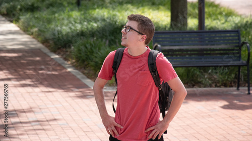 A young male student with thick black rimmed glasses and short hair wears a bright red shirt and black backpack stares up at what might be going on in the trees above him with his hands on his hips. © Joshua