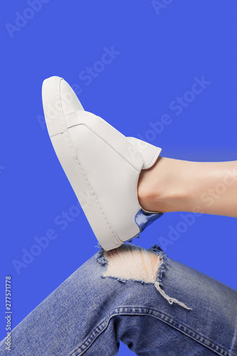 Trendy white eco leather sheakers on bent leg knee in ragged jeans over pastel purple blue background. Colorful main trend backdrop for your magazine, online store and media. Copy space