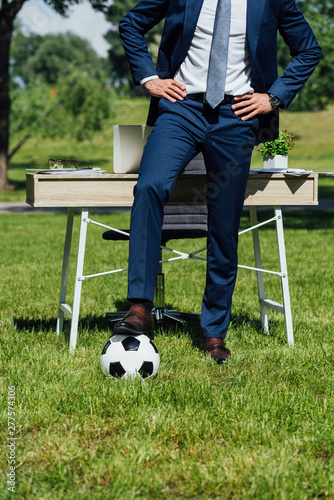 Cropped view of businessman standing with leg on soccer ball near table in park with hands on hips