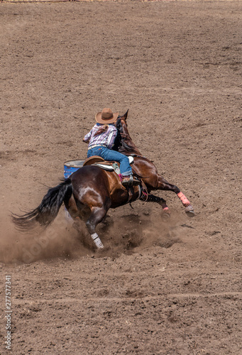 Barrel Racing at a Rodeo, cowgirl riding a dark colored horse around a  barrel. She has a pony tail and is wearing a large tan cowboy hat. The dirt  is flying as