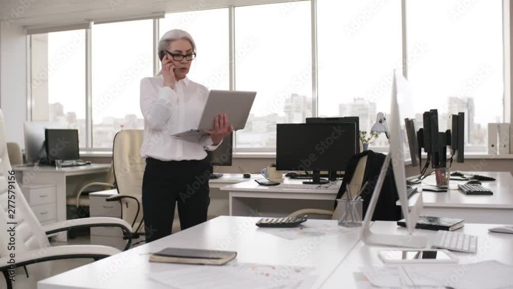 Medium shot of Caucasian businesswoman in her 50s, smartly dressed, with glasses and greying hair, walking around empty office with laptop, talking on mobile phone and looking at computer screen