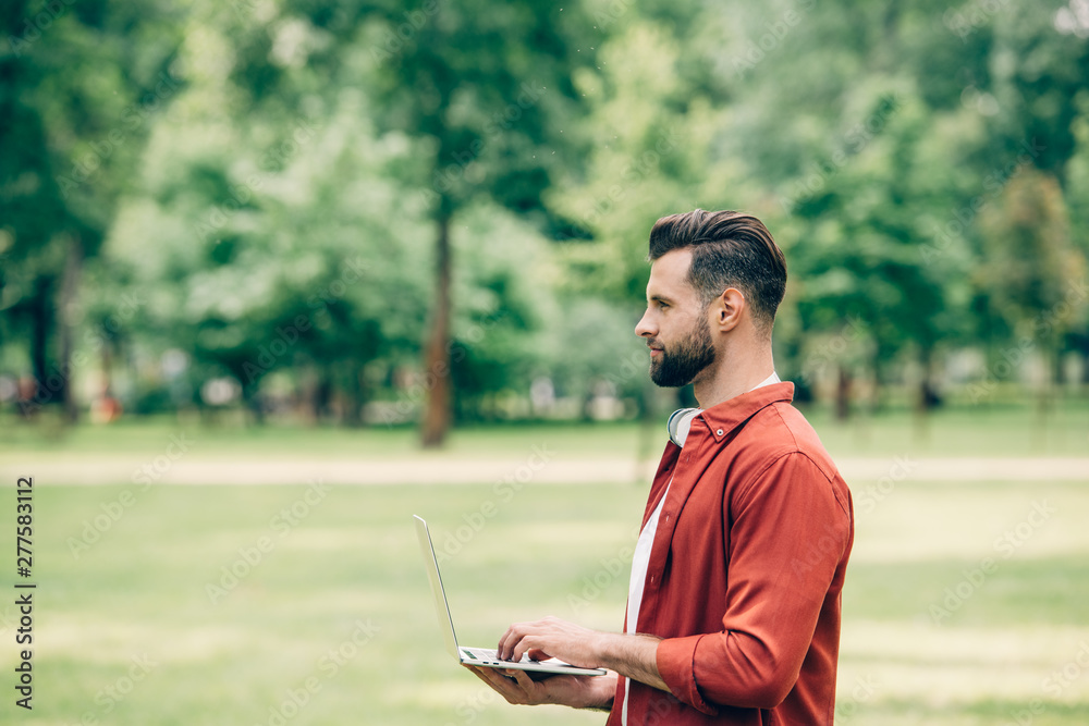 Obraz premium Side view of young man standing in park with laptop in hands and looking away