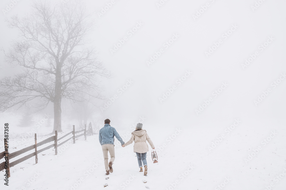 Rear of the young Caucasian couple in love holding hands and running during a blizzard in the countryside and girl holding a lantern. Back view. Rear of the young Caucasian couple in love holding hands and running during a blizzard in the countryside and girl holding a lantern. Back view.