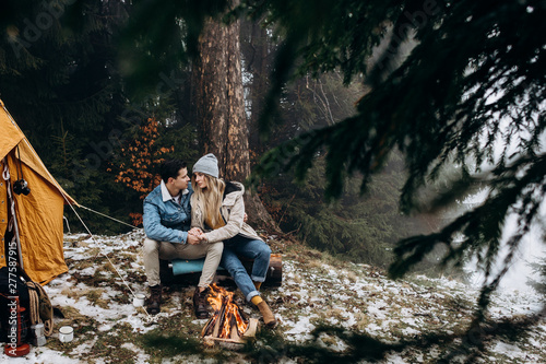 Portrait shot of th eyoungCaucasian couple sitting and huging at the tent in the middle of the forest in cold season during trip to mountains.