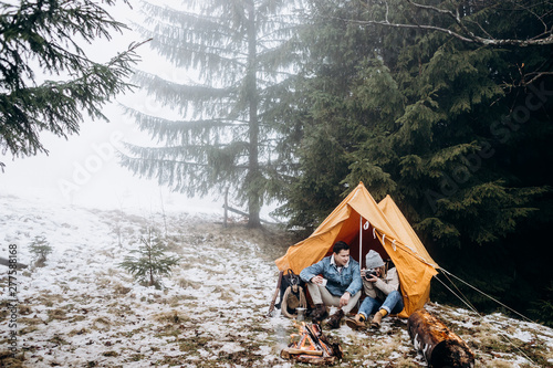 Vászonkép Young couple of travellers sitting at the yellow tent at the fire in the misty frosty forest on the cold day, man holding a cup of coffe and woman taking photos of the burning fire with a photocamera