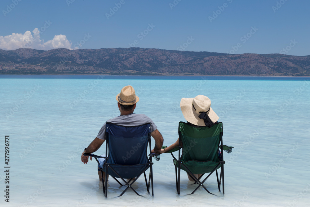 the beautiful girl and handsome man wearing wicker hats and sitting camp chairs in the lake and looking at the Salda lake from Turkey.