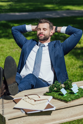 young businessman relaxing while sitting at table with sun batteries layout, glasses and noteboks, crossed lags and put hands behind head photo