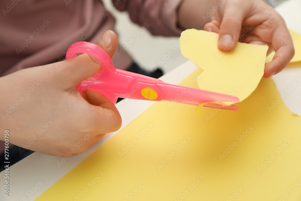 Child cutting out paper heart with plastic scissors at table, closeup