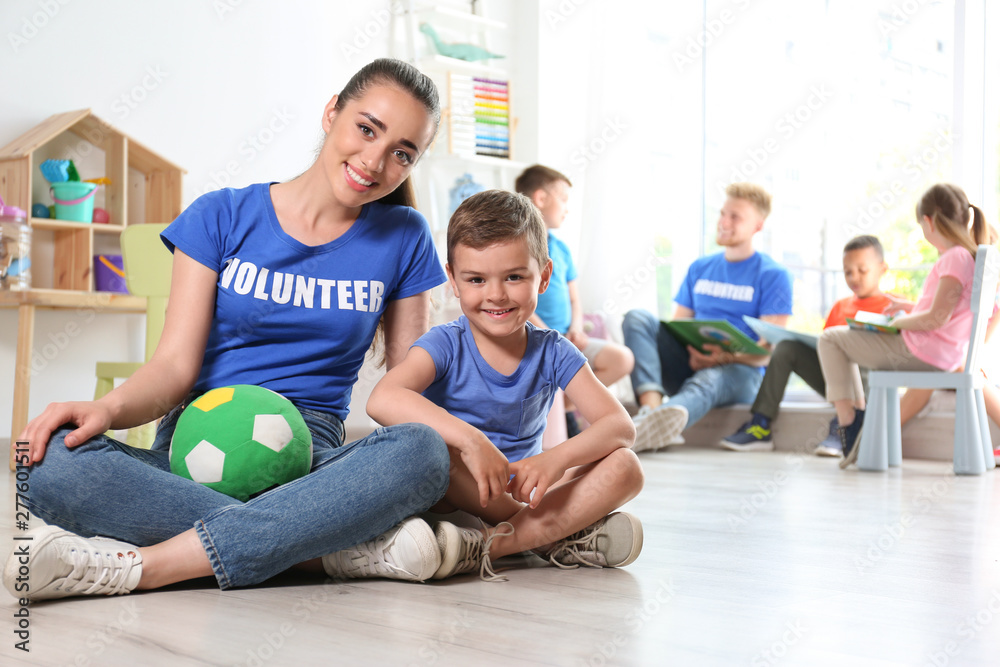 Young female volunteer with little boy sitting on floor indoors Young female volunteer with little boy sitting on floor indoors