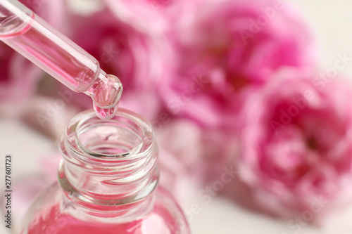 Fototapeta Naklejka Na Ścianę i Meble -  Dripping rose essential oil into bottle against blurred flowers on table, space for text