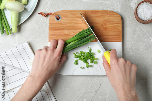Woman cutting fresh green onion on wooden board at marble table, top view