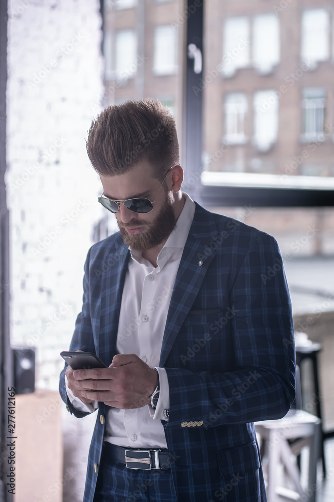 Vertical shot. Important message. Handsome young man in full suit holding a smart phone by standing at home Vertical shot. Important message. Handsome young man in full suit holding a smart phone by standing at home
