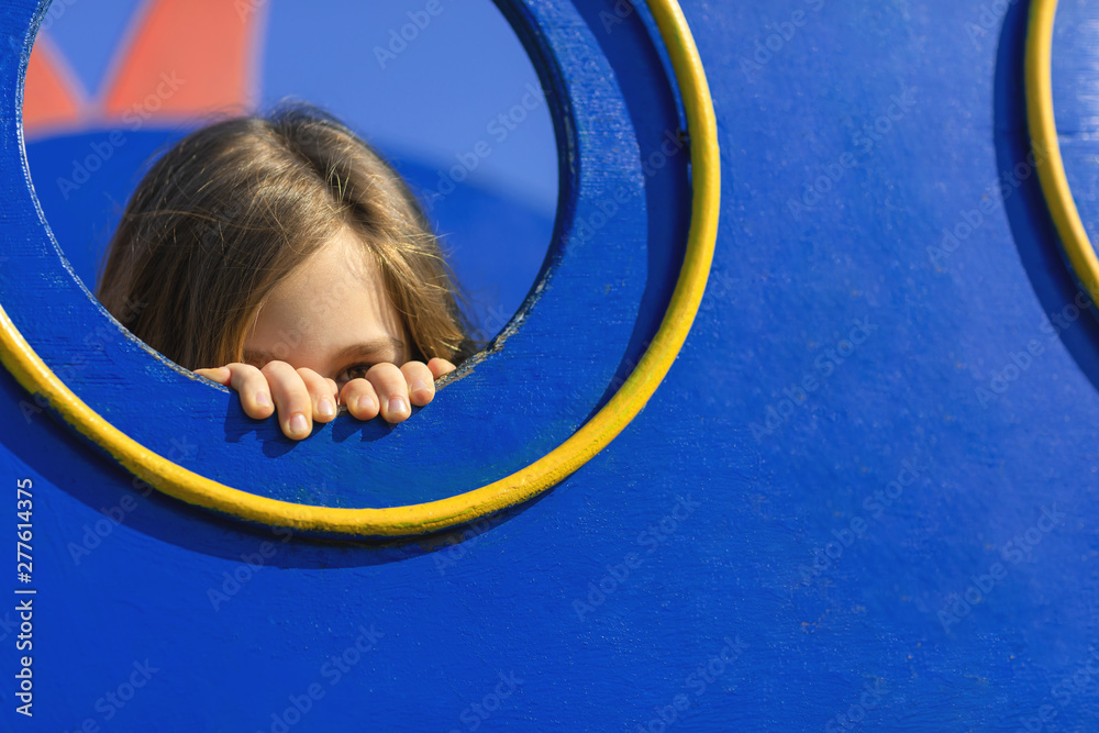 Young girl hiding and peeking behind hole in the wall of playhouse. Shy girl at playground. Scared kid concept. Young girl hiding and peeking behind hole in the wall of playhouse. Shy girl at playground. Scared kid concept.