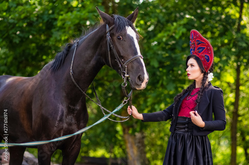 Lovely Fashion Model in Russian Style Kokoshnik Holding Thoroughbred Horse Closely.Posing Against Nature Background.