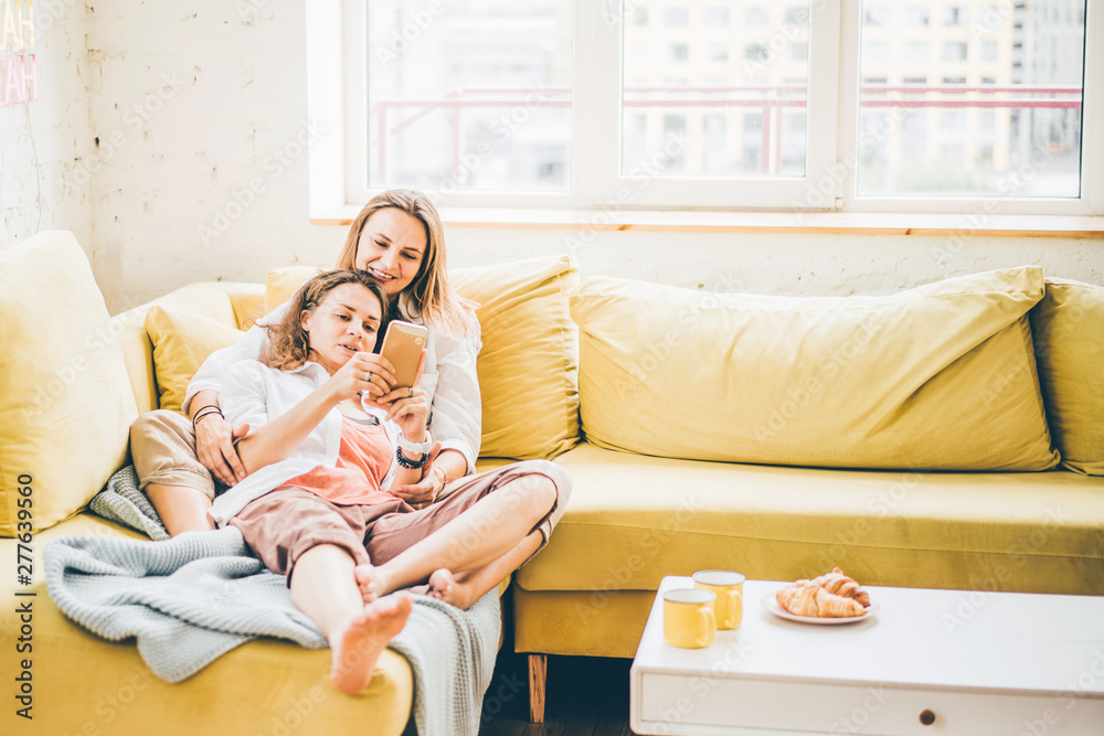 Girlfriends relaxing on colorful sofa in modern apartment. Girls looking on smartphone, smiling, embracing and enjoying togetherness. Concept LGBT couple at home. Girlfriends relaxing on colorful sofa in modern apartment. Girls looking on smartphone, smiling, embracing and enjoying togetherness. Concept LGBT couple at home.