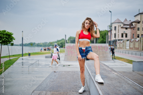 Sexy curly model girl in red top, jeans denim shorts, eyeglasses and sneakers posed at skatepark.