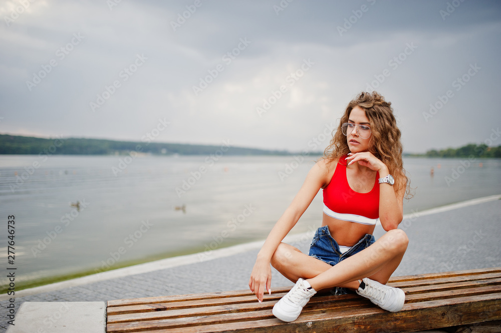 Sexy curly model girl in red top, jeans denim shorts, eyeglasses and sneakers posed on bench against lake.