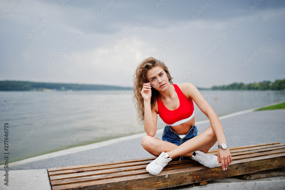 Sexy curly model girl in red top, jeans denim shorts, eyeglasses and sneakers posed on bench against lake.