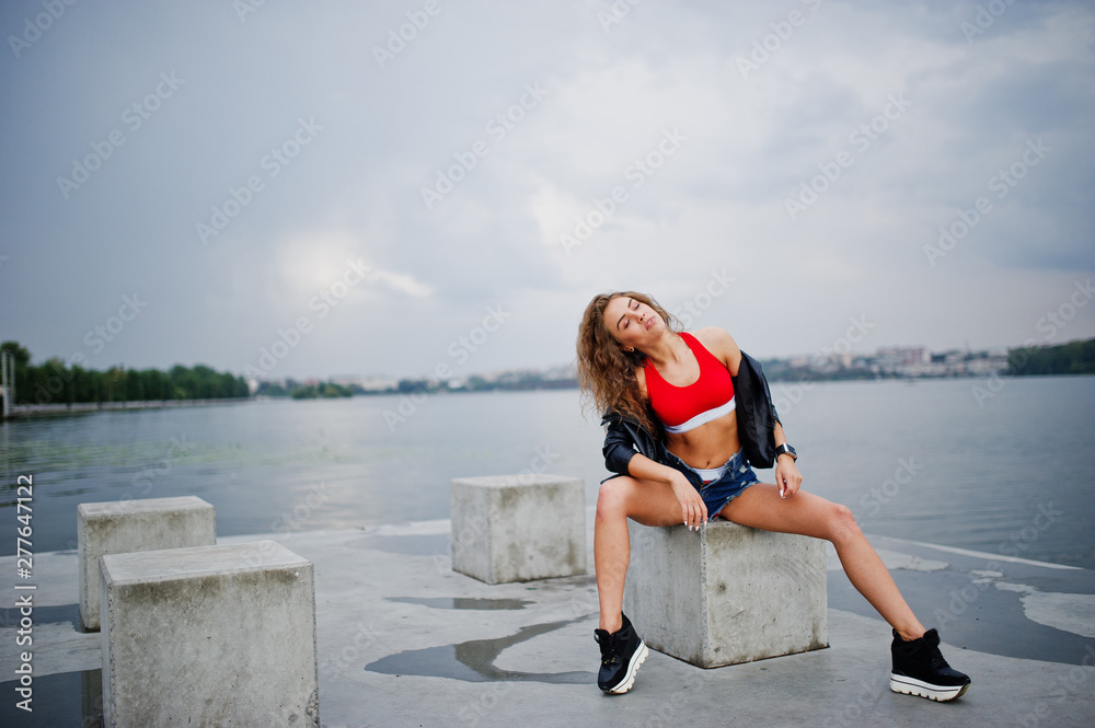 Sexy curly model girl in red top, jeans denim shorts, leather jacket and sneakers posed at stone cubes. Sexy curly model girl in red top, jeans denim shorts, leather jacket and sneakers posed at stone cubes.
