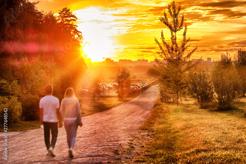 sunset at city park with couple walking along path towards setting sun and town skyline with dramatic sky background landscape street view of people enjoying summer evening authentic lifestyle scene