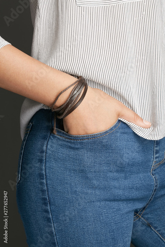 Cropped from top and bottom view shot of a woman with a bracelet on her arm. The fashion model on the gray background is dressed in a light striped shirt and jeans. You can see a multi-layer bracelet 