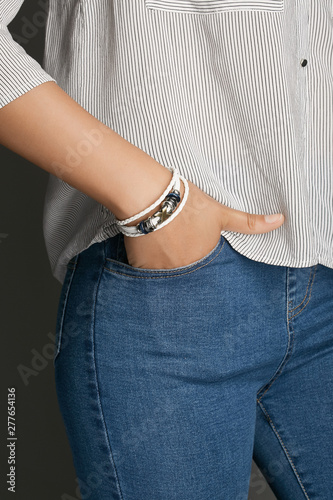 Cropped top and bottom photo of a woman with a bracelet on her arm. The model on a gray background is dressed in a light striped shirt and jeans. You can see a multi-layer bracelet made from a leather