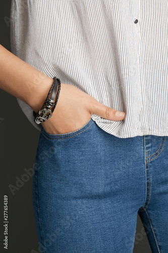 Cropped from top and bottom view shot of a woman with a bracelet on her arm. The fashion model on the gray background is dressed in a light striped shirt and jeans. You can see a multi-layer bracelet 