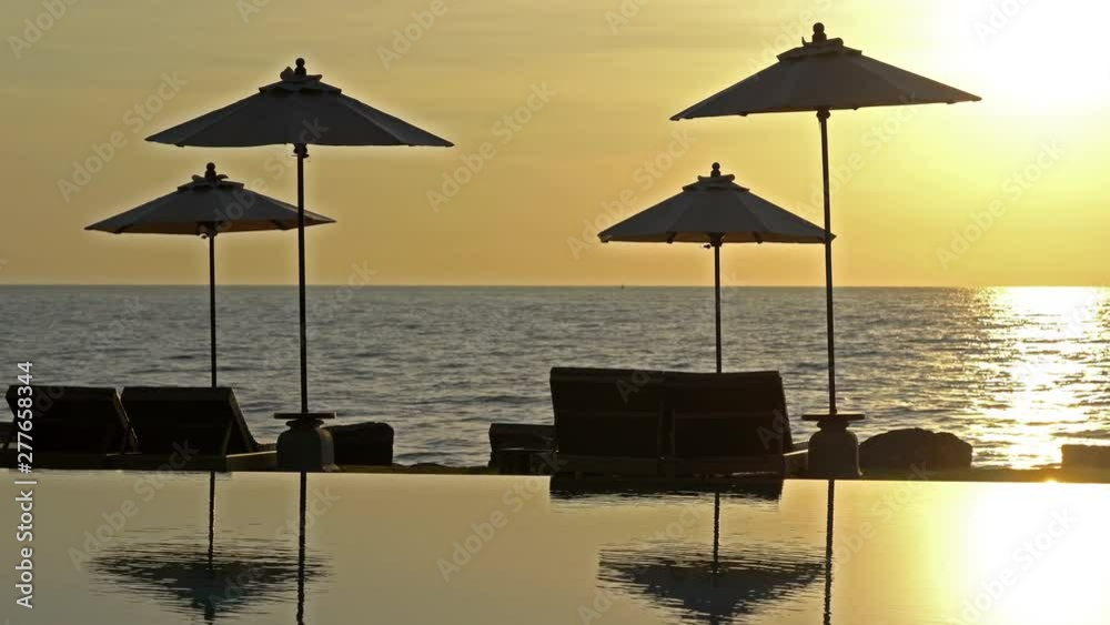 Static shot of beach sunbed loungers and umbrellas on the beach behind an infinity pool during sunset Static shot of beach sunbed loungers and umbrellas on the beach behind an infinity pool during sunset