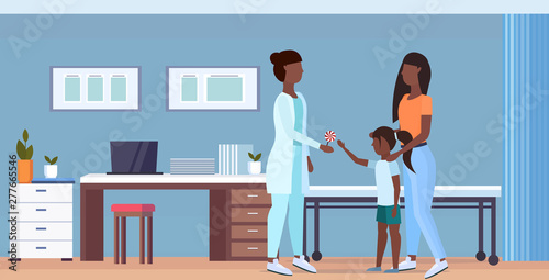 mother with daughter visiting african american doctor pediatrician giving lollipop to little girl consultation healthcare concept modern hospital interior full length flat horizontal