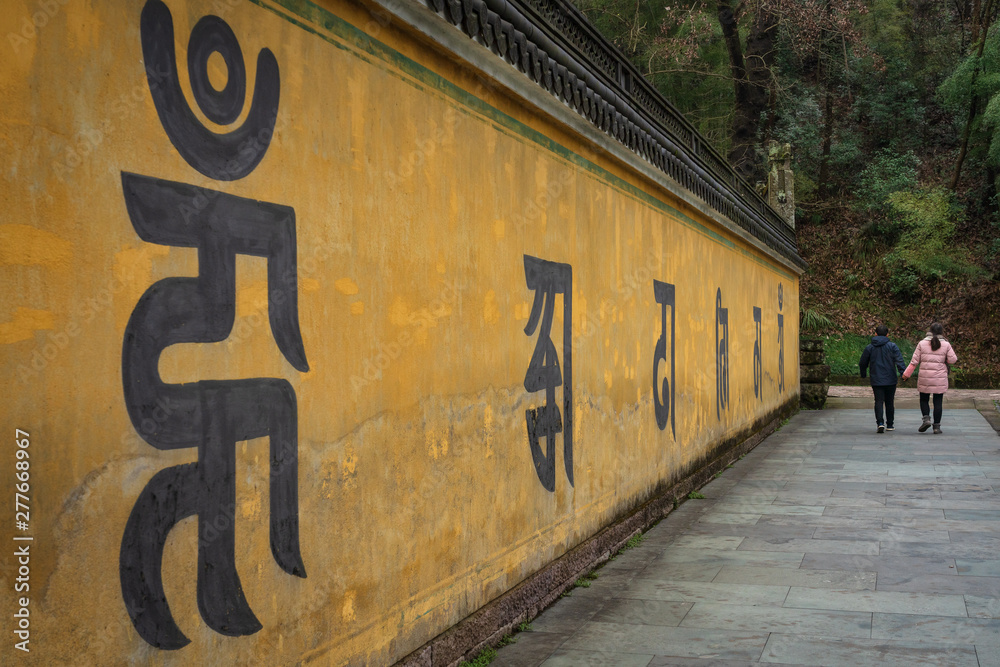 Great Buddha temple area, Xinchang, China - January 6, 2019 : Visitors of the temple complex walking past a long wall with inscriptions