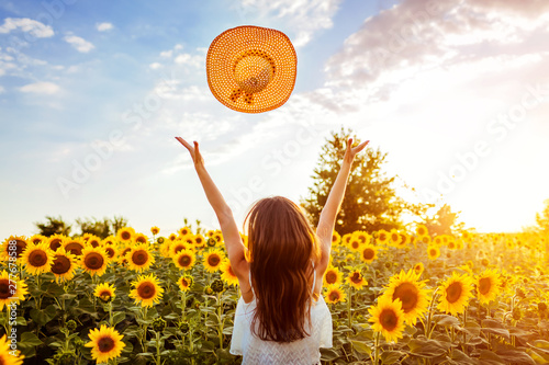 Young woman walking in blooming sunflower field throwing hat up and having fun. Summer vacation