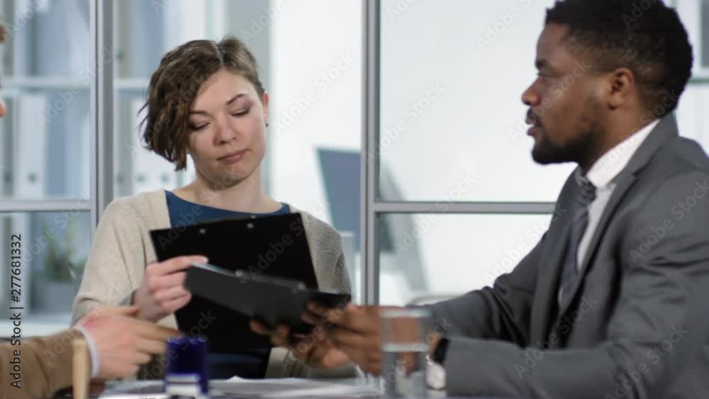 Tracking waist-up shot of Afro-American broker consulting young Caucasian client couple in company office, giving them draft copies of home insurance policy and explaining terms and conditions