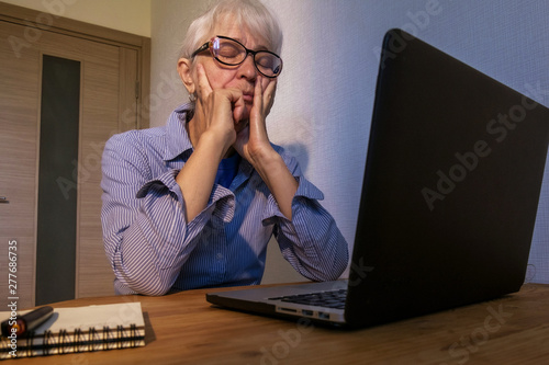 An elderly Caucasian woman in a blue and white shirt and blue T-shirt sadly closed her eyes and pressed her palms to her cheeks. Fear, confusion, depression