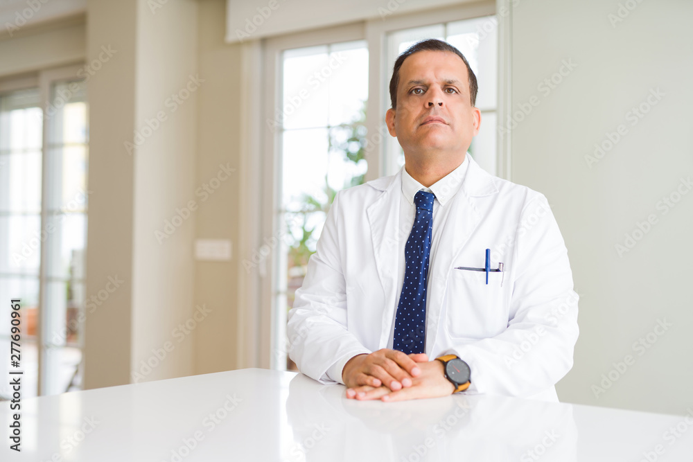 Middle age doctor man wearing medical coat at the clinic Relaxed with serious expression on face. Simple and natural looking at the camera.