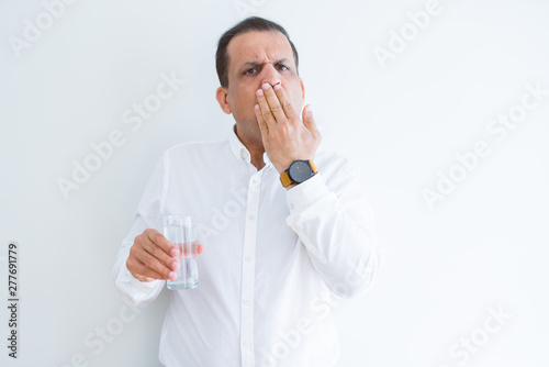 Middle age man drinking a glass of water over white background cover mouth with hand shocked with shame for mistake, expression of fear, scared in silence, secret concept
