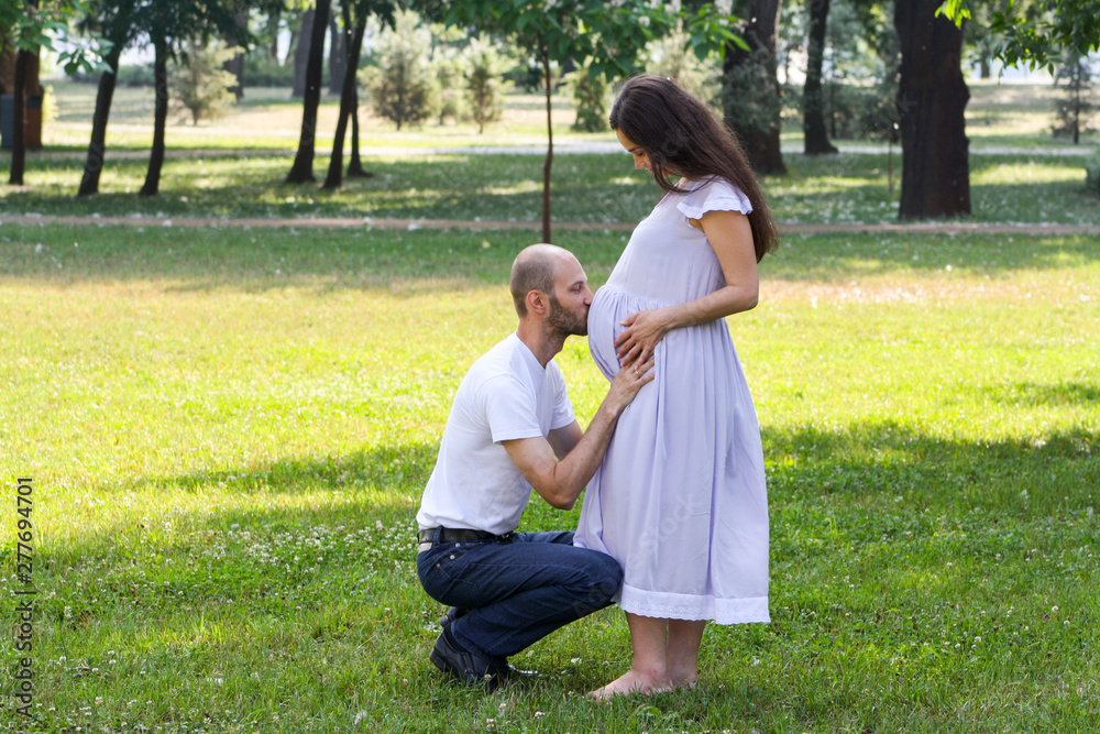 Portrait Of Husband Kissing His Wifes Big Pregnant Belly In The Park 