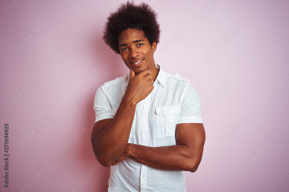 Young american man with afro hair wearing white shirt standing over isolated pink background looking confident at the camera with smile with crossed arms and hand raised on chin. Thinking positive. Young american man with afro hair wearing white shirt standing over isolated pink background looking confident at the camera with smile with crossed arms and hand raised on chin. Thinking positive.
