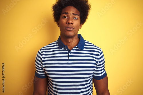 American man with afro hair wearing navy striped polo standing over isolated yellow background with serious expression on face. Simple and natural looking at the camera.