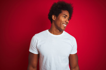Young american man with afro hair wearing white t-shirt standing over isolated red background looking away to side with smile on face, natural expression. Laughing confident.